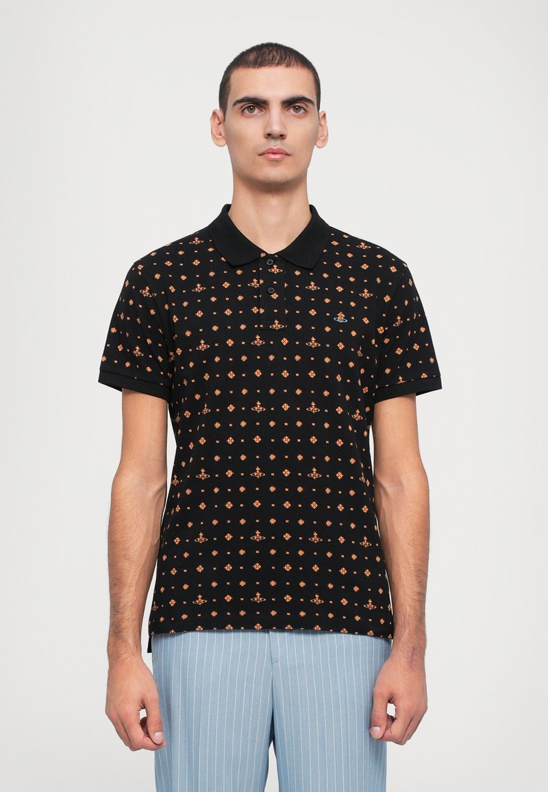 Μαύρο πόλο μπλουζάκι με πορτοκαλί γεωμετρικό μοτίβο, κοντά μανίκια και μαύρο γιακά, φορεμένο με φωτεινές μπλε ριγέ παντελόνες.