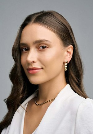 Orecchini a goccia dorati con tre perle testurizzate, abbinati a una collana in oro, indossati con una camicia bianca con colletto su uno sfondo grigio.