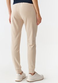 Beige joggingbyxor med smalnande ben, elastiska muddar och två bakfickor; har en broderad björnlogga ovanför den högra fickan.