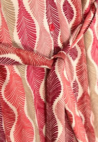 Vêtement en coton avec un motif de vagues dans des nuances de rouge, rose et beige, avec un bouton central rouge et un détail de taille nouée.