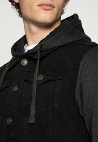 Svart denimjacka med grå huva, med en knappstängning fram, två fickor och en strukturerad yta. Dragsnören hänger från huven.