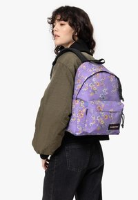 Zaino floreale viola con spallacci regolabili, tasca frontale, cerniere nere e logo "EASTPAK". Indossato sopra un giaccone verde oliva.