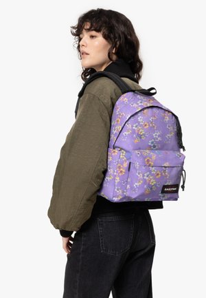 Lila blommig ryggsäck med justerbara axelremmar, framficka, svarta dragkedjor och "EASTPAK"-logotyp. Bärs över en olivgrön jacka.