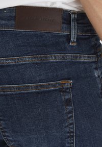 Sötétkék farmernadrág bőr folttal a deréknál, rajta a "CASUAL FRIDAY" felirattal, amely bele van nyomva. Figyelemre méltó varrási részletek a zsebek és varrások mentén.