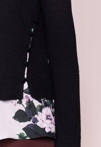 Černý pletený kardigan s texturovanou látkou, se zdobnými kulatými knoflíky po straně a košilí s květinovým vzorem viditelnou pod ním.