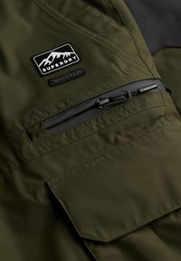 Olijfgroene jas van een gestructureerde stof, met een ritszak en een patch met een berglogo. Duurzaam ontwerp met versterkte stiksels.