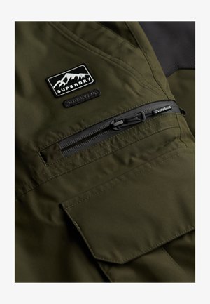 Olijfgroene jas van een gestructureerde stof, met een ritszak en een patch met een berglogo. Duurzaam ontwerp met versterkte stiksels.