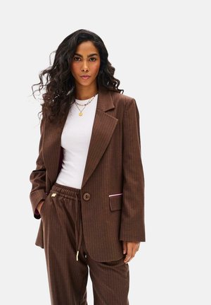 Kobieta o długich kręconych włosach, ubrana w brązową marynarkę w prążki i dopasowane spodnie, na białej bluzce, stoi z jedną ręką w kieszeni.
