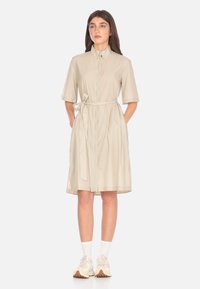 Abito camicia color beige chiaro con maniche corte, chiusura frontale con bottoni e vita legata. Presenta una silhouette rilassata e arriva poco sopra il ginocchio.