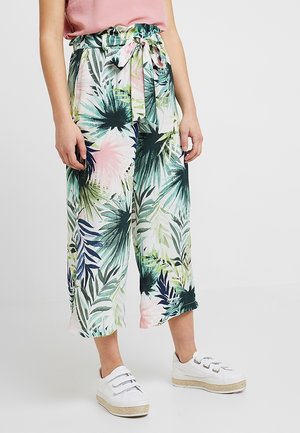 Nainen, jolla on leveälahkeiset trooppikuvioiset housut, joissa on solmittava vyötärönauha, ja valkoiset tarrakiinnitteiset tennarit, seisomassa yksiväristä taustaa vasten.