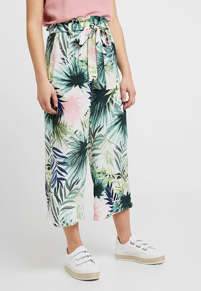 Femme portant un pantalon à jambes larges à imprimé tropical avec une ceinture nouée et des baskets blanches à scratch, debout devant un fond uni.