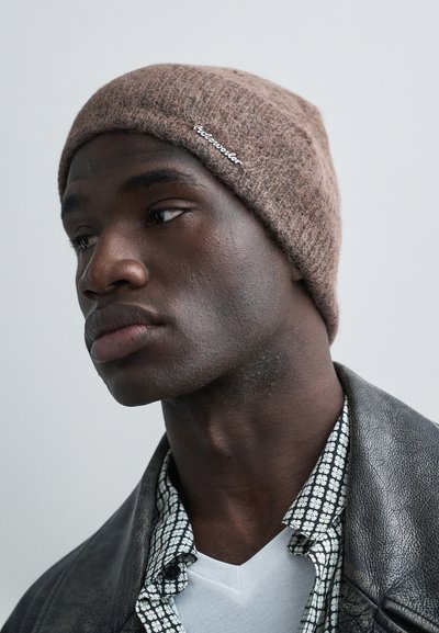 Rudas megztas kepurė su minkštu, tekstūruotu paviršiumi, puošta balta logotipu. Dėvima asmens, vilkinčio juodą odinę striukę, margintą marškinius ir baltą marškinėlį.