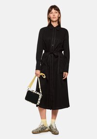 Schwarzes Hemdkleid mit Taillengürtel, zwei vorderen Taschen und kontrastierenden Nähten. Kombiniert mit einer gestreiften Handtasche und gemusterten Sneakers.