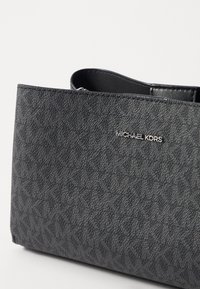 Sac à main texturé gris avec un motif géométrique, doté d'une plaque logo en métal et d'une simple anse supérieure. Le matériau semble être du simili cuir.