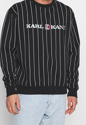 Schwarzer Langarmpullover mit weißen vertikalen Streifen und dem Schriftzug "KARL KANI" auf der Brust, kombiniert mit hellblauen Jeans.