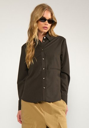 Femme blonde portant une chemise noire à fines rayures boutonnée, un pantalon beige et des lunettes de soleil cat-eye noires sur un fond clair uni.
