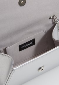 Clutch de imitación de cuero plateado con textura suave, cierre metálico a presión y bolsillo interior. Presenta una etiqueta negra con la marca "even&odd".