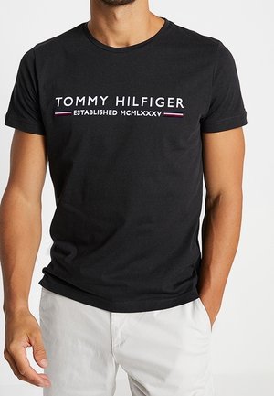 Mand iført en sort Tommy Hilfiger t-shirt med hvid tekst og romertal, kombineret med lyse bukser, hænderne afslappet langs siden.