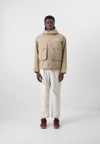 Beige jacket with a hood, front zipper, and two large chest pockets. Worn over light-colored pants and brown sneakers with white soles.