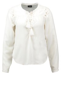 Blouse blanche en tissu léger avec des manches longues, des accents en dentelle sur les épaules et un lien de fermeture avec des franges au niveau du col.