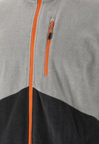 Grå zip-up jakke med tekstureret stof, med en orange lynlås og et sort nederste panel, der skaber et farveblok design.
