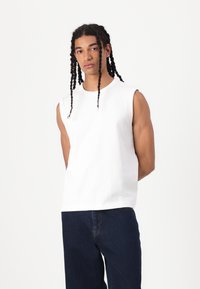 Tricou alb fără mâneci din bumbac, cu gât rotund, textură netedă și croială lejeră, asortat cu blugi din denim de culoare albastru închis.