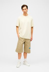 Joven de pie con las manos en los bolsillos, vistiendo una camiseta beige de gran tamaño, pantalones cortos caqui hasta la rodilla con un logo azul, calcetines blancos y zapatillas deportivas blancas.