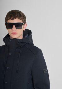 Marineblauwe hooded jacket met een gladde textuur, drukknoopsluiting en trekkoorden. Bevat een logopatch op de mouw en wordt gecombineerd met oversized zwarte zonnebril.