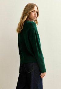 Maglione lavorato a maglia di colore verde scuro, con una vestibilità ampia, colletto rotondo e polsini a costine, abbinato a pantaloni in denim scuro a vita alta.