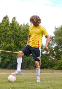 Gul og rød atletiktrøje med Adidas branding, sorte shorts og livlige fodboldstøvler. Fodbold på græsset med mål i baggrunden.