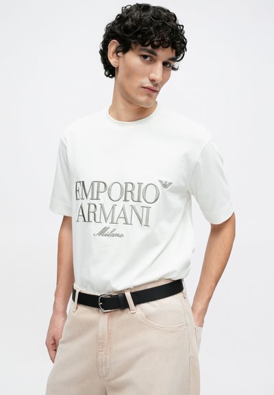 Biele bavlnené tričko s nápisom "EMPORIO ARMANI Milano" vytlačeným v šedej farbe. Kombinované so svetlobéžovými nohavicami a čiernym opaskom. Viditeľné kučeravé vlasy.