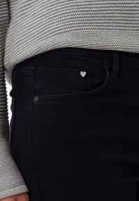 Jeans de mezclilla oscura con corte slim, que presentan un adorno en forma de corazón plateado en el bolsillo delantero. Suéter de punto gris texturizado visible encima.