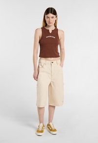 Cropped top senza maniche marrone con logo, pantaloni cargo beige con tasche laterali e sneakers in suede gialle con lacci bianchi. Texture lisce.