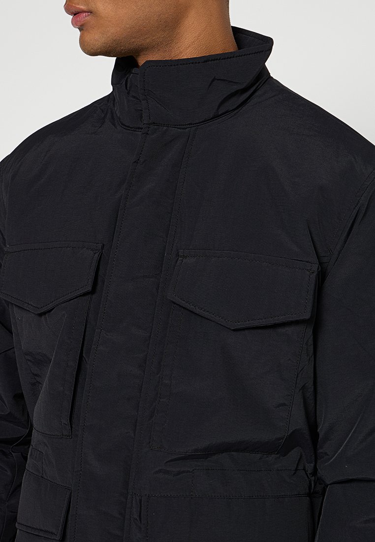 Giacca nera isolata con colletto alto, numerose ampie tasche frontali e una texture liscia e elegante. Dettagli di cucitura robusta sono visibili.