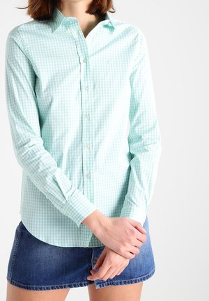 Woman wearing a light green checkered button-up shirt and a blue denim skirt, standing with hands clasped in front.