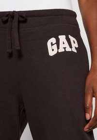 Pantalones de chándal marrón oscuro con cintura ajustable, que presentan el logotipo bordado en blanco "GAP" en el muslo izquierdo. Textura suave, diseño casual.