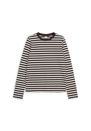 Femme portant un long manteau beige, un pull bleu marine drapé sur les épaules, une chemise rayée, un jean bleu, un sac à main noir et des sandales noires à talons.