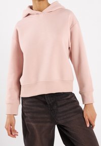 Sweat à capuche en coton rose à coupe décontractée, avec capuche à cordon et ourlet côtelé. Associé à un jean foncé présentant une texture subtile.