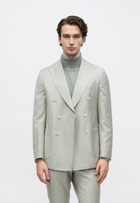 Ung mand med bølget hår iført lysegrå, dobbeltradet jakkesæt med nålestriber og grå rullekrave, stående mod en ensfarvet hvid baggrund.