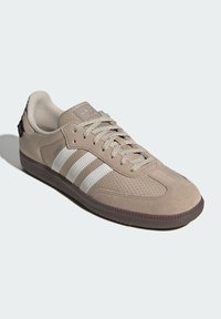 Beige Adidas sneaker with white stripes, textured fabric, suede panels, tan laces, and a brown rubber sole, shown from a side front angle.