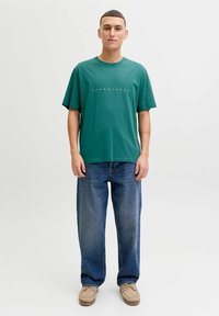 Groen katoenen T-shirt met korte mouwen en een ronde halslijn, met witte tekst "JACK & JONES." Gecombineerd met losse blauwe spijkerbroek en beige schoenen.