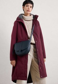Bordeauxjas met een grijze fleece-voering bij de kraag, gecombineerd met een kabelgebreide crèmekleurige trui en beige corduroybroek. Zwarte crossbodytas.