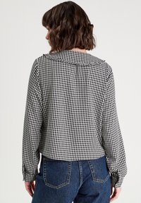 Schwarz-weiße karierte Bluse mit Kragen und langen Ärmeln, im cropped Design und mit knöpfbaren Manschetten. Kombiniert mit hochgeschnittenen blauen Jeans.