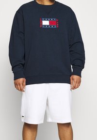 Granatowa bluza z okrągłym dekoltem i długimi rękawami, z haftowanym logo w kolorach czerwonym, białym i niebieskim. Noszona z białymi szortami.