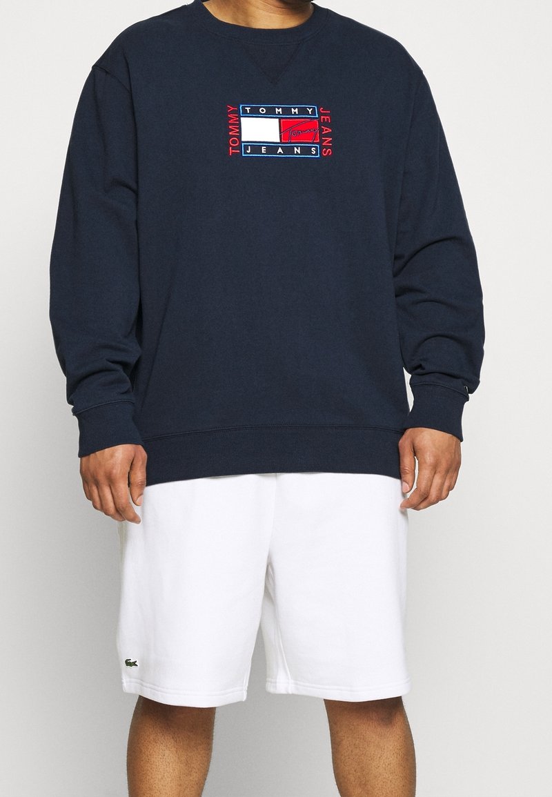 Granatowa bluza z okrągłym dekoltem i długimi rękawami, z haftowanym logo w kolorach czerwonym, białym i niebieskim. Noszona z białymi szortami.