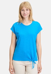Niebieska bluzka z krótkim rękawem, z teksturowanym wzorem, okrągłym dekoltem i regulowanym dołem ze sznurkiem. Wykonana z lekkiego materiału.