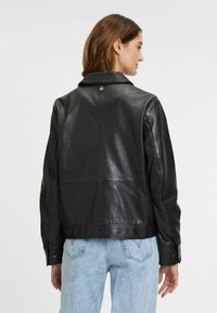 Schwarze Lederjacke mit klassischem Kragen, Knopfdetails und kurzem Schnitt, kombiniert mit hellblauen Jeans.