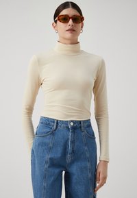 Beige prodloužený top s dlouhým rukávem a stojáčkem vyrobený z pružného materiálu, v kombinaci s modrými džíny s vysokým pasem. Hladká struktura a přiléhavý střih.
