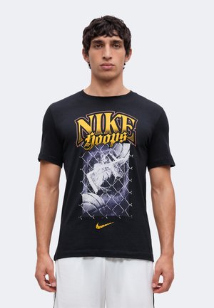 Άνδρας που φοράει μαύρη μπλούζα Nike Hoops με γραφικό μπάσκετ και λευκή βερμούδα, στέκεται μπροστά σε απλό φόντο.