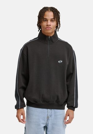 Sweat-shirt zippé noir avec col montant, présentant des bandes blanches sur les côtés et un logo sur la poitrine. Tissu doux, coupe ample, ourlet côtelé.